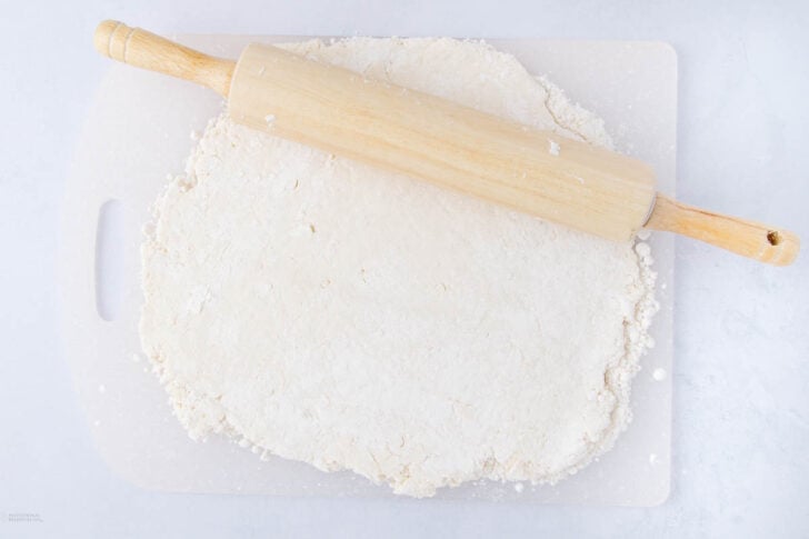 A wooden rolling pin rests on top of a slab of dough being rolled out on a white cutting board, with flour scattered around.