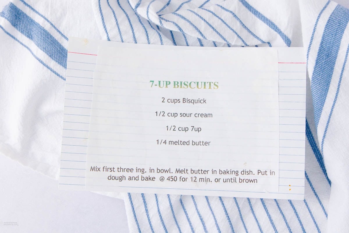 A recipe card titled "7-UP BISCUITS" lists ingredients and instructions, placed on a white cloth with blue stripes. The card includes Bisquick, sour cream, 7UP, and melted butter, with baking instructions at the bottom.