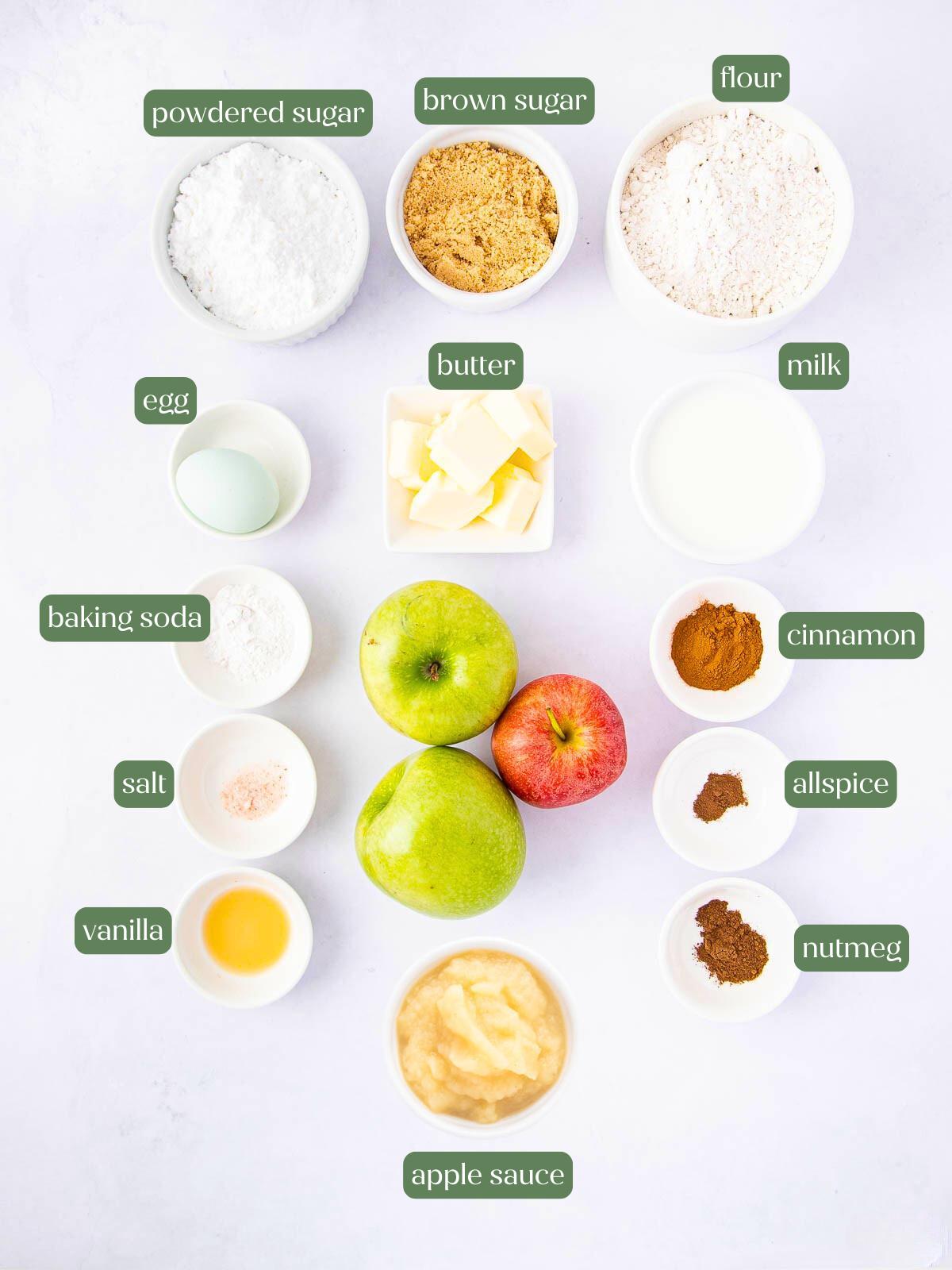 Flat lay of labeled baking ingredients in bowls and cups, including powdered sugar, brown sugar, flour, egg, butter, milk, baking soda, cinnamon, allspice, salt, vanilla, nutmeg, three apples, and applesauce.