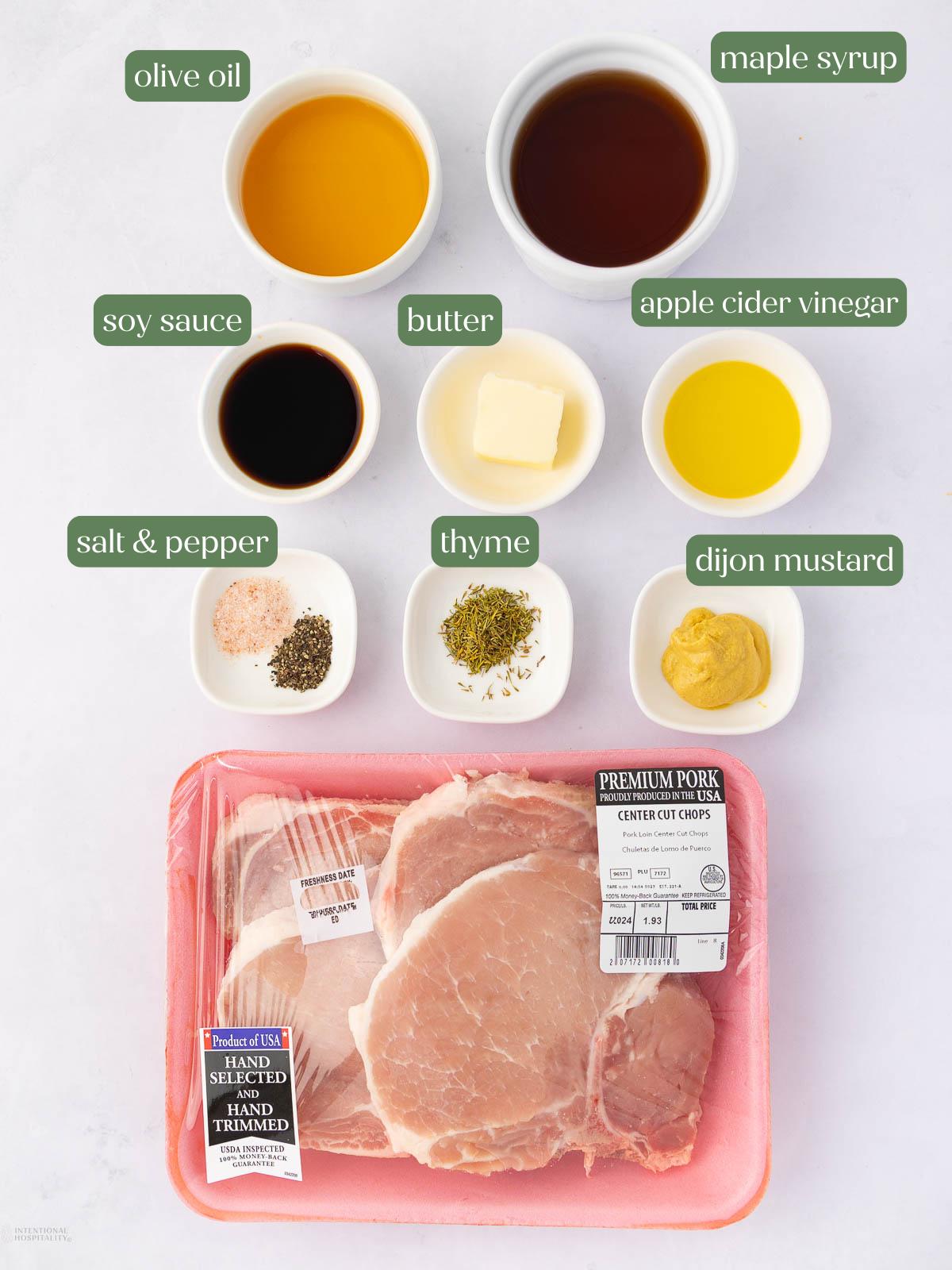 Ingredients for a pork chop recipe arranged on a white background: raw pork chops, olive oil, maple syrup, soy sauce, butter, apple cider vinegar, salt and pepper, thyme, and dijon mustard in small bowls.