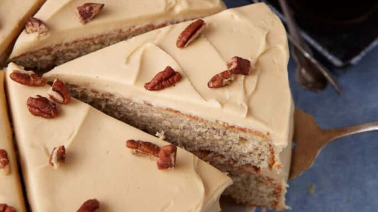 A slice of frosted layer cake being lifted from the whole cake. The cake has light brown icing and is topped with pecan halves.