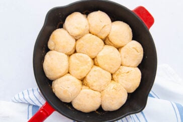 A cast iron skillet filled with golden, freshly baked biscuits sits on a white surface with a blue-striped kitchen towel underneath.