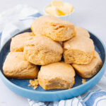 A blue plate filled with golden brown, fluffy biscuits stacked on top of each other, with a small bowl of butter in the background and a striped cloth underneath the plate.