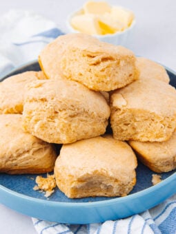 A blue plate filled with golden brown, fluffy biscuits stacked on top of each other, with a small bowl of butter in the background and a striped cloth underneath the plate.