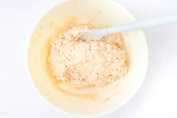 A white mixing bowl with sticky, beige dough being stirred by a light blue spatula, set on a white surface.