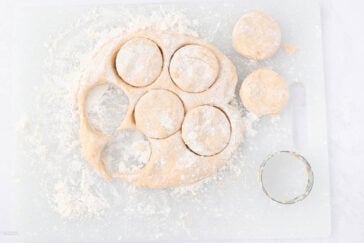A floured surface with biscuit dough and several biscuits cut out using a round cutter, leaving circular shapes in the dough. Some biscuits are placed nearby, ready to be baked.