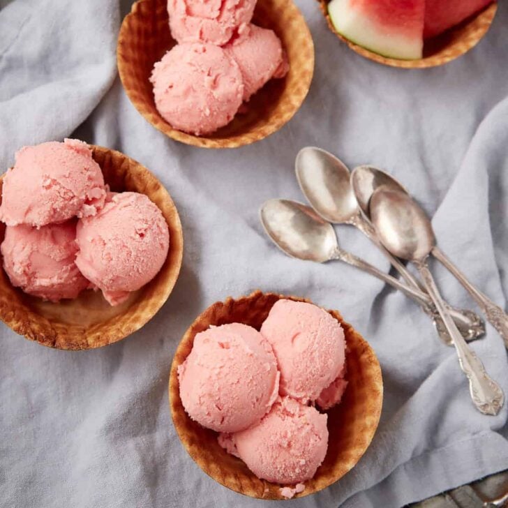 Four wooden bowls filled with scoops of pink ice cream are on a light cloth, next to four silver spoons and a slice of watermelon.