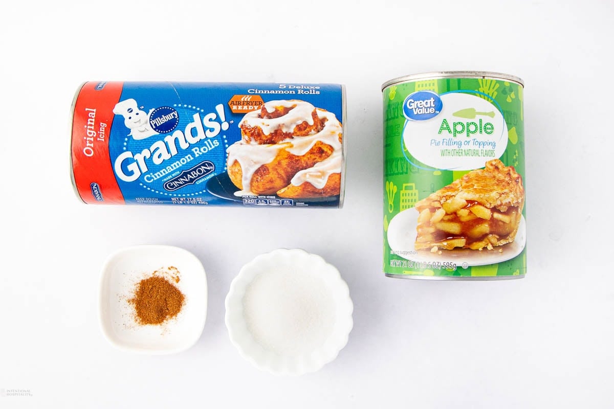 A can of Pillsbury Grands! cinnamon rolls, a can of Great Value apple pie filling, a small dish of cinnamon, and a small dish of sugar arranged on a white background.