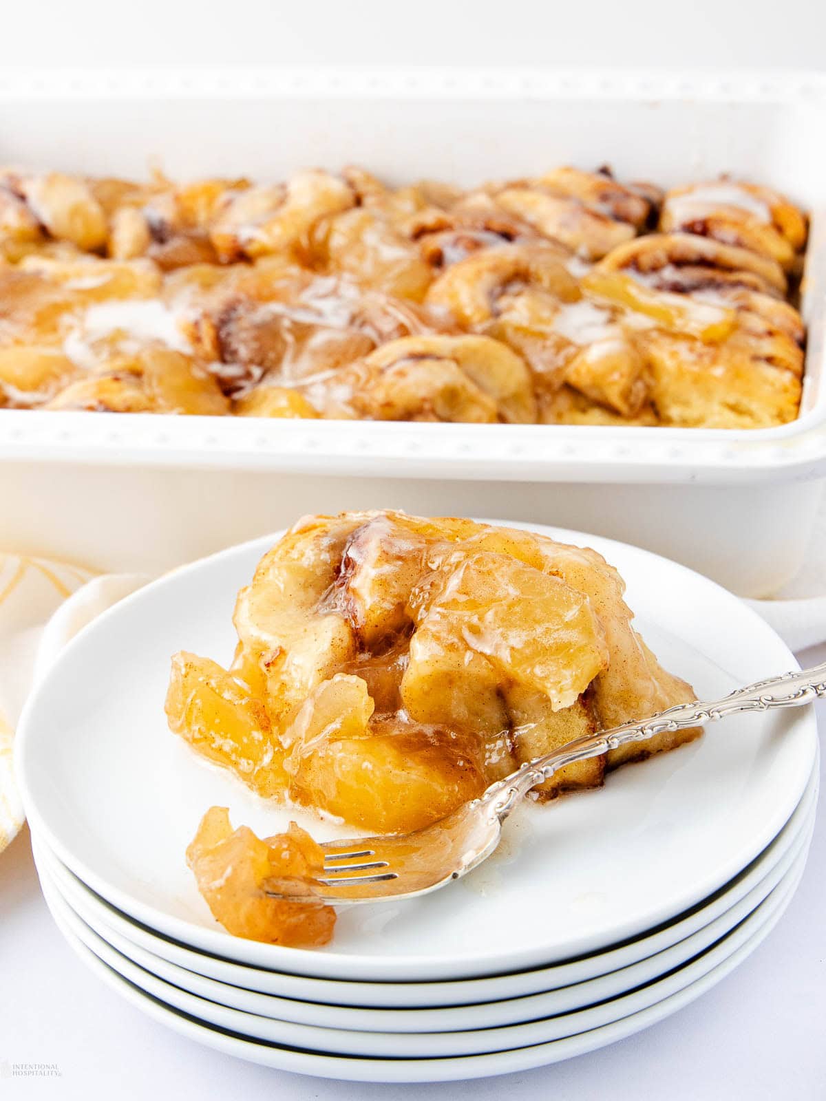 A serving of warm peach cobbler with golden, flaky crust and peaches sits on a white plate with a fork. The rest of the cobbler is in a white baking dish in the background.