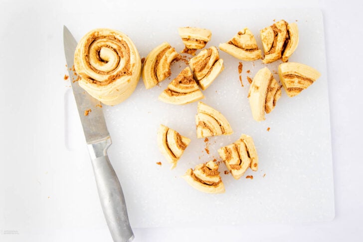 A large cinnamon roll being sliced into smaller pieces on a white cutting board with a silver knife beside it. Crumbs are scattered around the board.