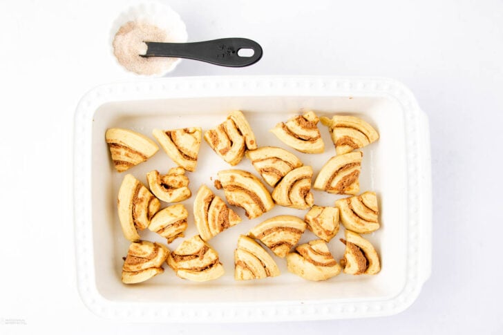 White baking dish filled with cut pieces of cinnamon rolls, arranged in a single layer. Above the dish is a black measuring spoon containing a mixture of cinnamon and sugar on a white background.