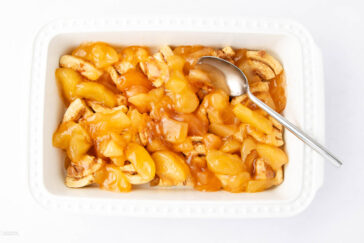 A white rectangular dish filled with cinnamon roll pieces topped with glossy apple pie filling. A metal spoon rests in the dish on the right side.