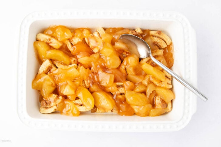 A white rectangular dish filled with cinnamon roll pieces topped with glossy apple pie filling. A metal spoon rests in the dish on the right side.