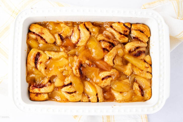 A white rectangular baking dish filled with a golden brown cobbler, featuring swirled pieces of baked dough and glossy, syrupy apple filling. A yellow and white striped towel is partially visible underneath.