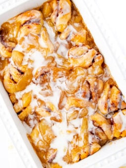 A white baking dish filled with cinnamon roll pieces coated in a gooey, golden caramel sauce and drizzled with white icing. The rolls appear soft and freshly baked.