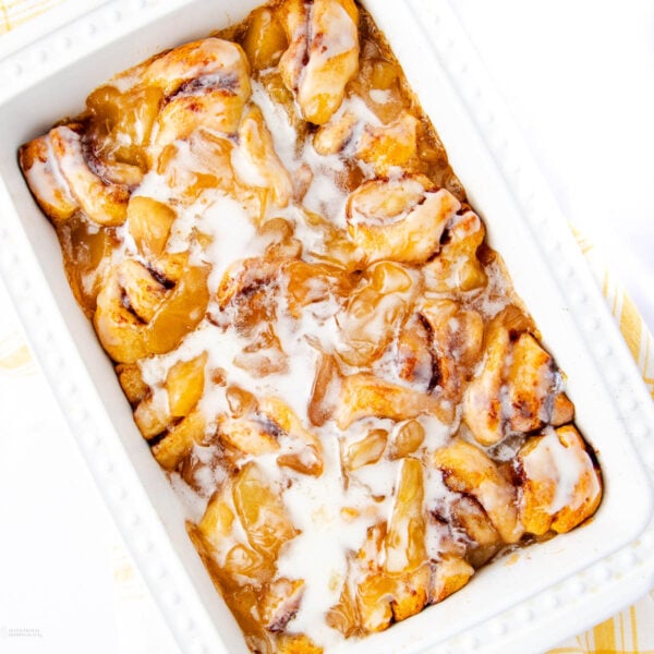 A white baking dish filled with cinnamon roll pieces coated in a gooey, golden caramel sauce and drizzled with white icing. The rolls appear soft and freshly baked.
