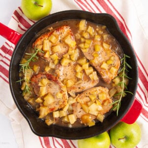 A skillet with four pork chops topped with diced apples and sauce, garnished with rosemary sprigs. The skillet sits on a red-and-white striped towel with green apples nearby.