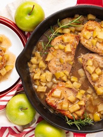 A skillet filled with pork chops topped with diced apple sauce sits next to a plate with a serving of the dish. Fresh green apples, a red-striped towel, and rosemary garnish surround the skillet.