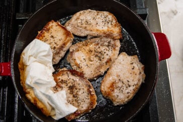 Five seasoned pork chops are cooking in a black cast iron skillet on a stovetop. A hand holding a paper towel is blotting excess oil from one of the chops.