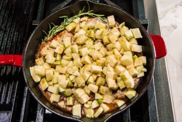A cast iron skillet on a stove filled with diced green apples, a sprig of rosemary, and browned meat simmering in sauce.