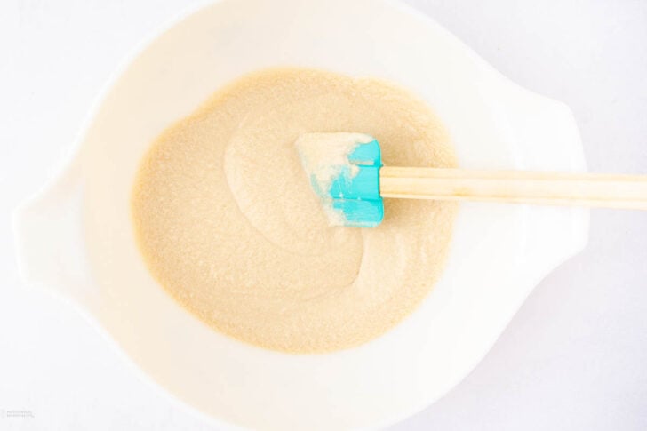 A white bowl with a light beige mixture inside, being stirred with a blue silicone spatula with a wooden handle. The background is white.