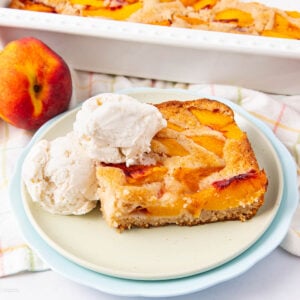 A slice of peach cobbler topped with two scoops of vanilla ice cream sits on a plate, with a whole peach and a baking dish of cobbler in the background.