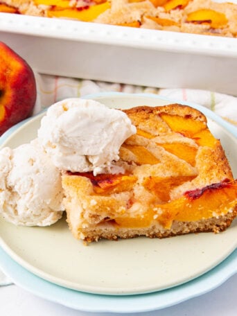 A slice of peach cobbler topped with two scoops of vanilla ice cream sits on a plate, with a whole peach and a baking dish of cobbler in the background.