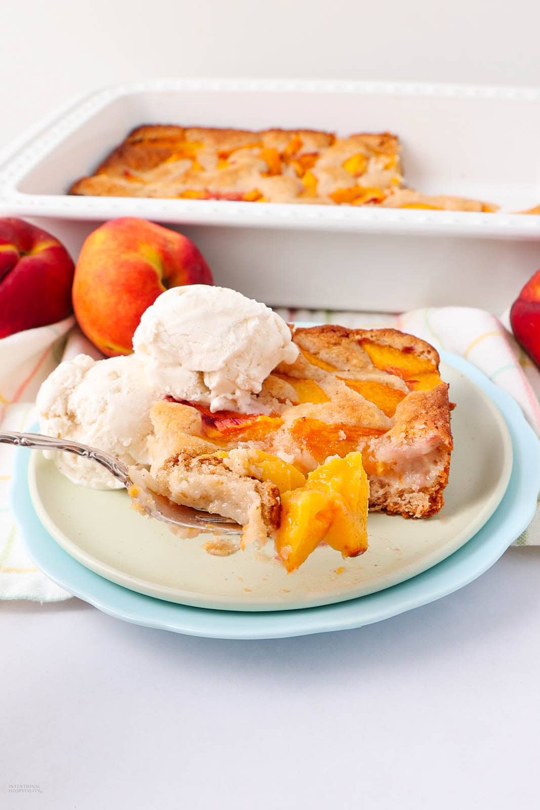 A slice of peach cobbler with vanilla ice cream on a plate, with fresh peaches and a baking dish of cobbler in the background. A fork rests on the plate, holding a bite of cobbler.