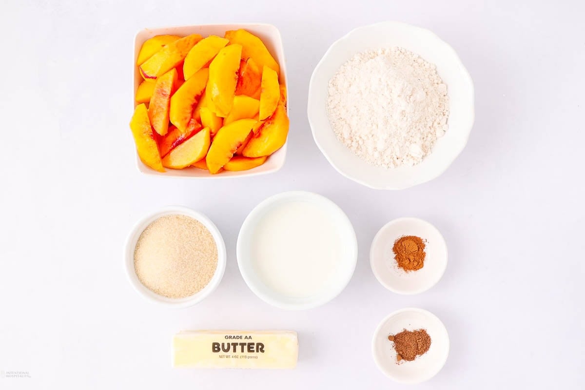 Six ingredients for a peach cobbler are arranged on a white surface: sliced peaches, flour, sugar, milk, a stick of butter, and two small bowls of spices (likely cinnamon and nutmeg).