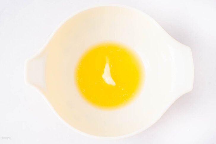 A white mixing bowl containing melted butter with a small lump of unmelted butter in the center, viewed from above on a white background.
