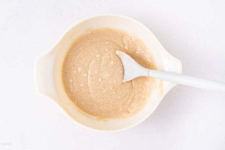 A white mixing bowl filled with light brown batter, with a white spatula resting inside, placed on a white surface.
