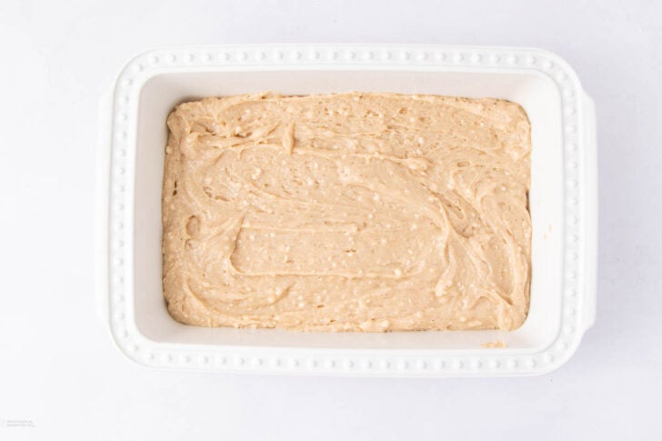A white rectangular baking dish filled with smooth, light tan batter, spread evenly and ready to be baked, sits on a white surface.