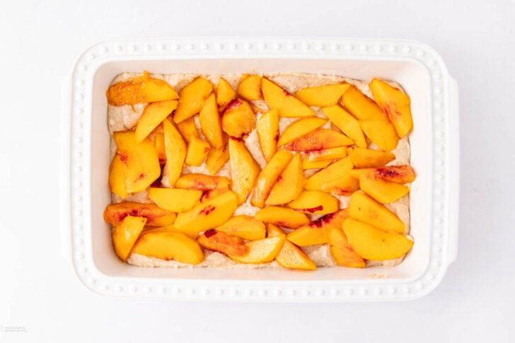 A white rectangular baking dish filled with an even layer of fresh, sliced peaches arranged over an unbaked batter on a white background.