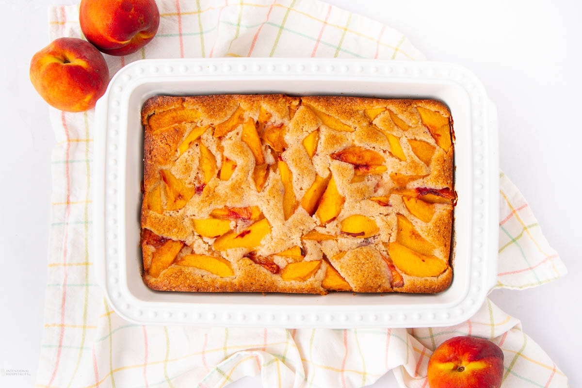 A rectangular baking dish with golden-brown peach cobbler sits on a plaid kitchen towel, surrounded by fresh peaches.