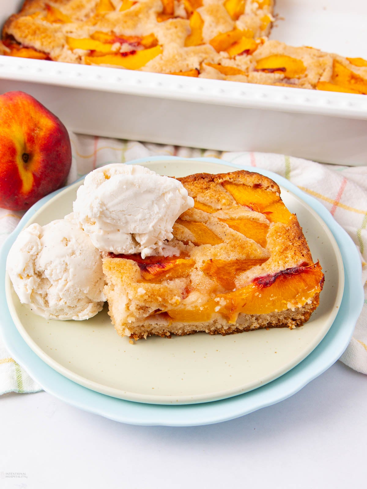 A square piece of peach cobbler with golden baked peach slices is served on a plate with two scoops of vanilla ice cream. A fresh peach and the baking dish are nearby in the background.