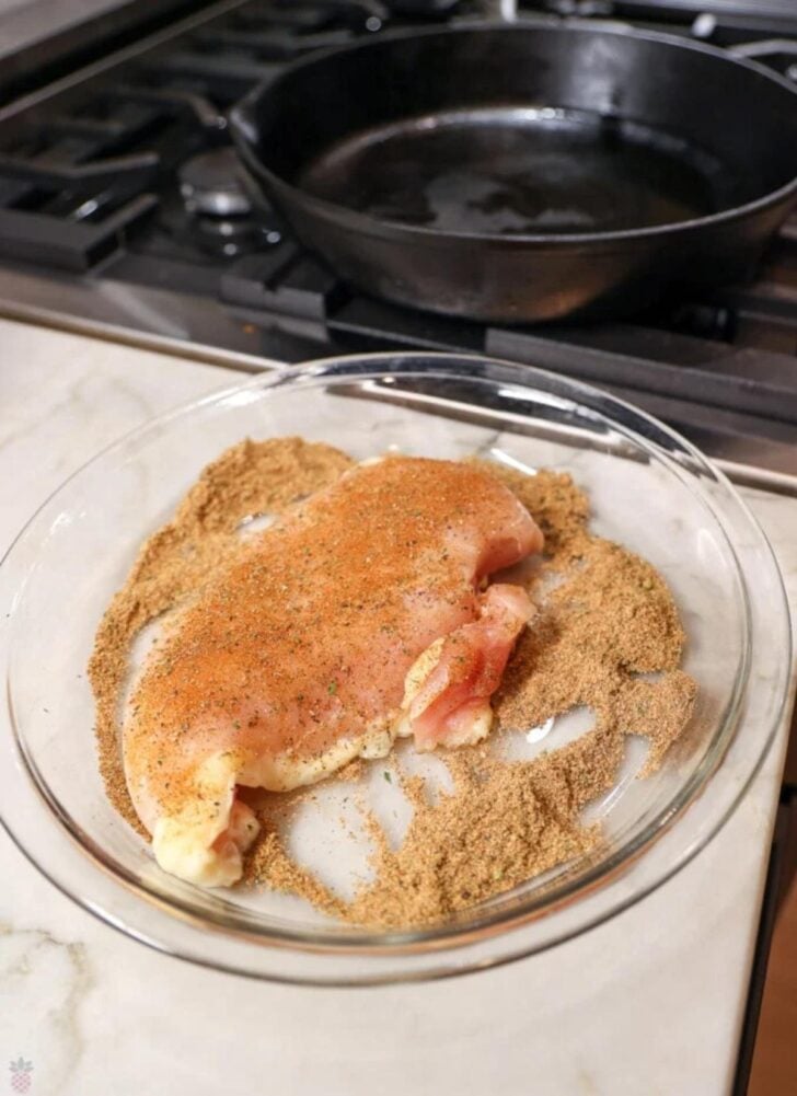 A raw, seasoned chicken breast sits on a bed of breadcrumbs in a glass dish, ready for coating. In the background, a black cast-iron skillet is heating on a stovetop.