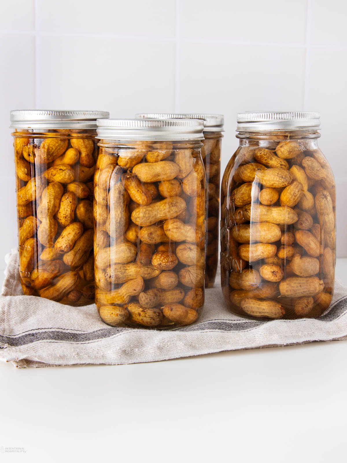 Four glass jars filled with boiled peanuts in brine are placed on a folded striped cloth, against a white tiled background.