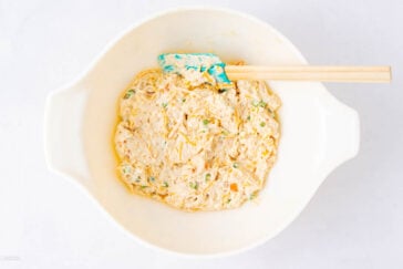 A white mixing bowl contains a creamy, chunky mixture with visible bits of green and orange, and a blue spatula with a wooden handle rests inside the bowl.