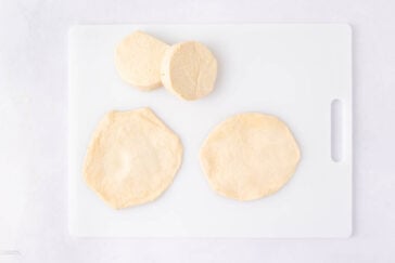 Two rounds of biscuit dough and two flattened discs of dough are on a white cutting board with a handle, set against a white background.
