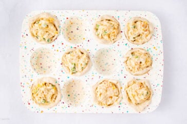 A muffin tin with colorful dots holds six raw biscuit dough cups filled with a creamy mixture, while three sections are empty. The filling contains visible vegetables.