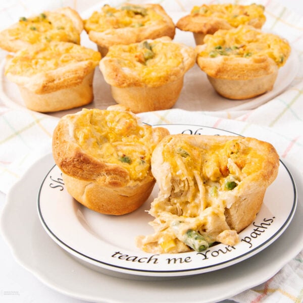 Mini chicken pot pies with golden, flaky crusts and cheesy tops are arranged on a plate and in the background. One pie is cut open, revealing a creamy filling with chicken and mixed vegetables.