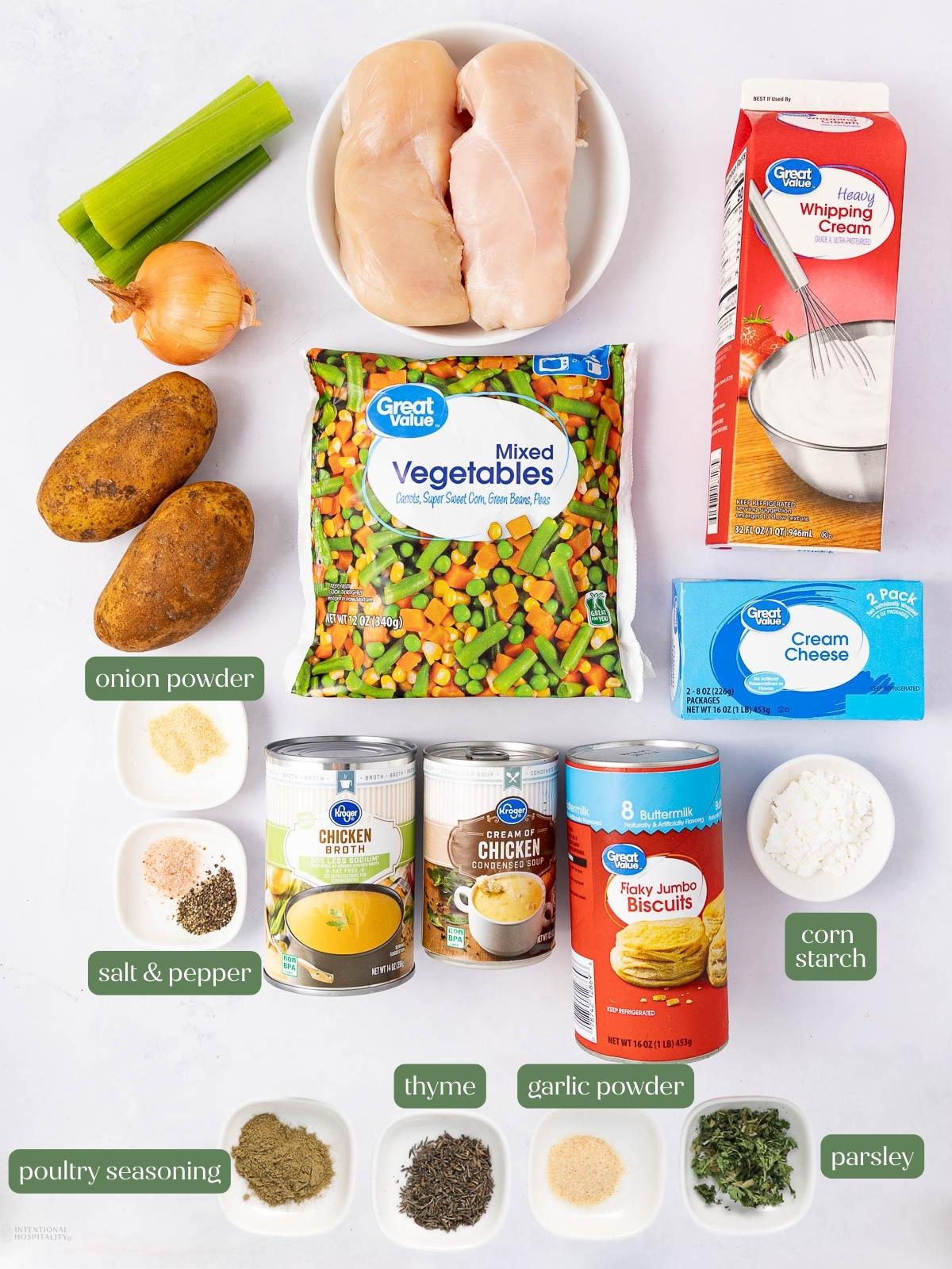 Ingredients for a chicken pot pie casserole are laid out on a white surface, including chicken breasts, potatoes, celery, mixed vegetables, cream cheese, whipping cream, biscuits, broth, and various spices in small bowls.