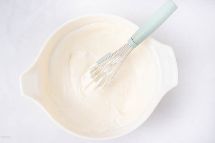 A white mixing bowl filled with creamy, whipped mixture and a light blue whisk resting inside on a white surface.