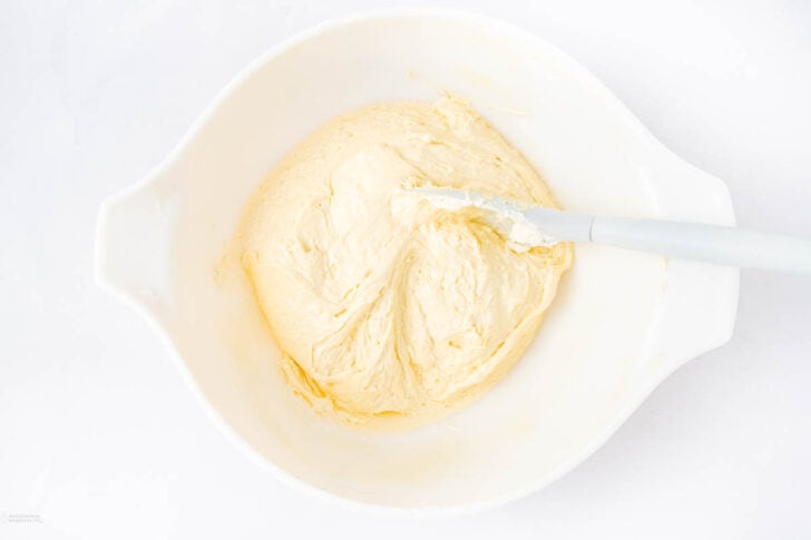 A white mixing bowl filled with creamy, pale yellow batter, with a white spatula resting inside, on a white surface.