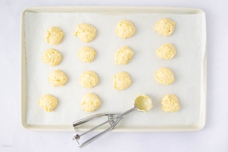 A baking sheet lined with parchment paper holds 16 evenly spaced dollops of raw cookie dough. A metal cookie scoop with dough residue rests on the corner of the tray.