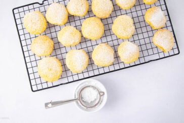 Yellow cookies topped with powdered sugar are arranged on a black cooling rack. Below the rack, a small sifter with powdered sugar rests on a white dish, all set against a white background.