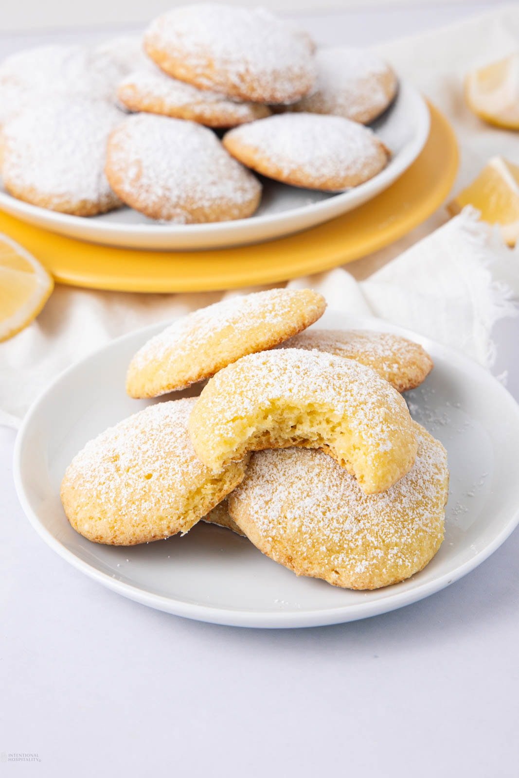 A plate of lemon cookies dusted with powdered sugar, with one cookie showing a bite taken out. Another plate of cookies and lemon wedges are in the background.