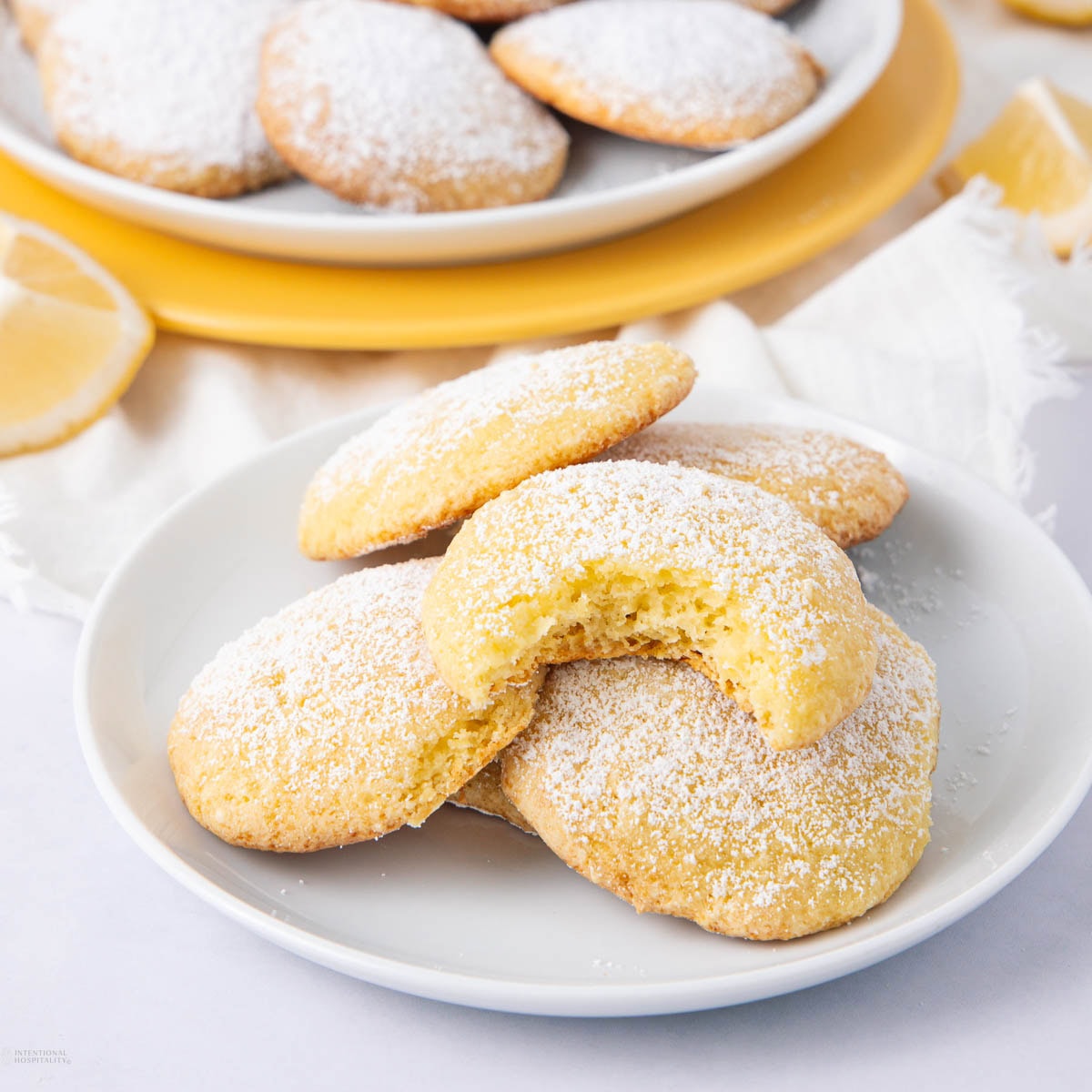 A plate of lemon cookies dusted with powdered sugar, with one cookie showing a bite taken out.