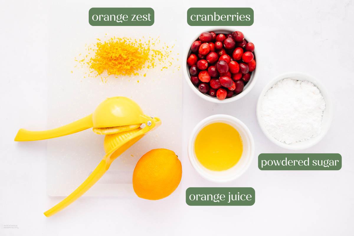 A cutting board with orange zest, fresh cranberries, powdered sugar, orange juice in a bowl, a halved orange, and a yellow citrus juicer arranged neatly on a white surface. Labels identify each ingredient.