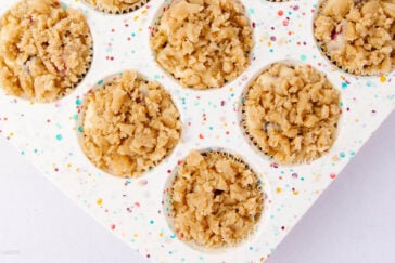 A close-up of a white muffin tin with colorful specks, filled with paper liners and unbaked muffins topped with crumbly streusel topping.